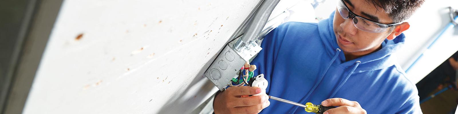 Student wearing safety googles and tool belt installing electrical switch on cement wall