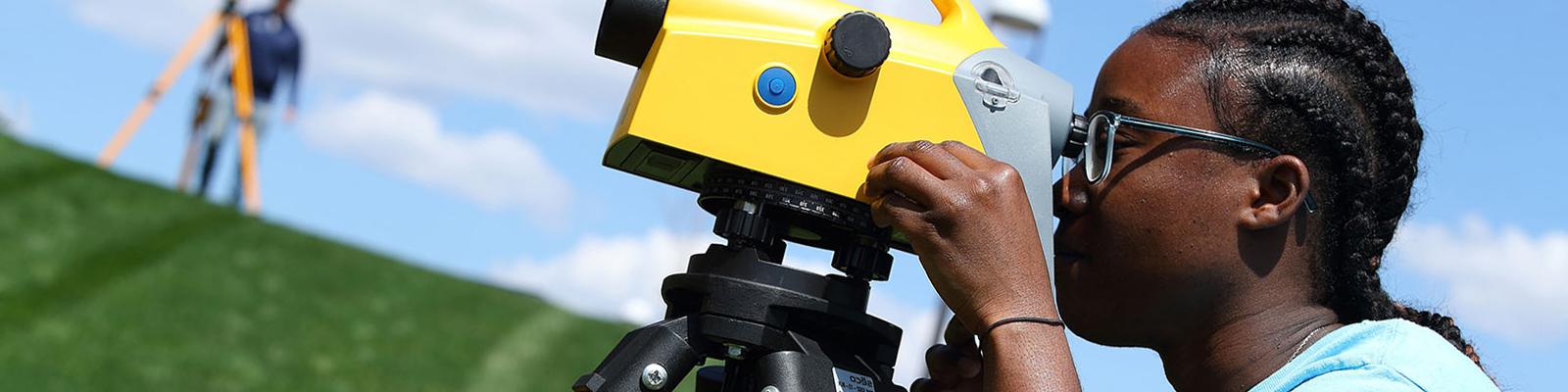 Woman at bottom of grassy hill, looking into surveying equipment. Soft focus background on another student.